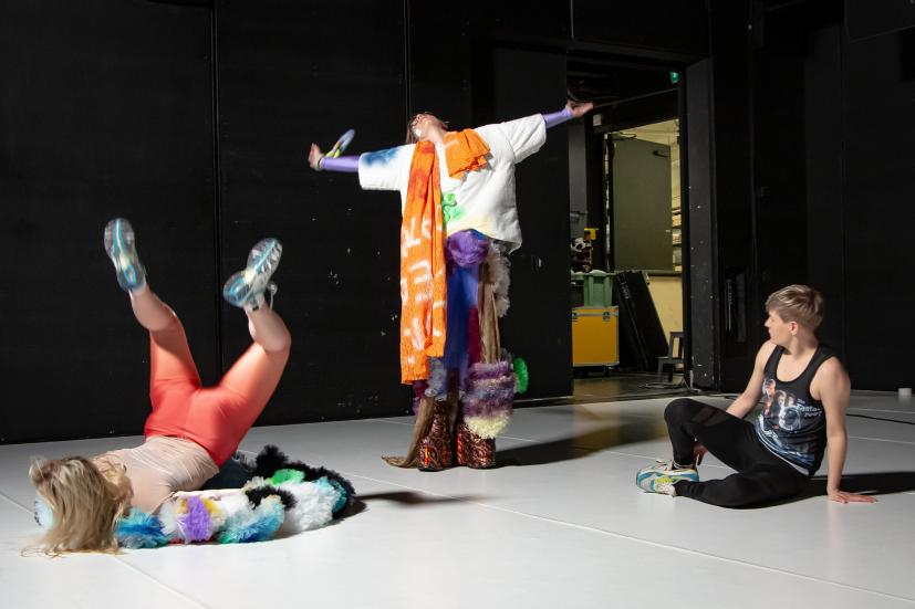 Three performers. One is lying on their stomach. One is standing with their hands spread out. The third is sitting on the ground.