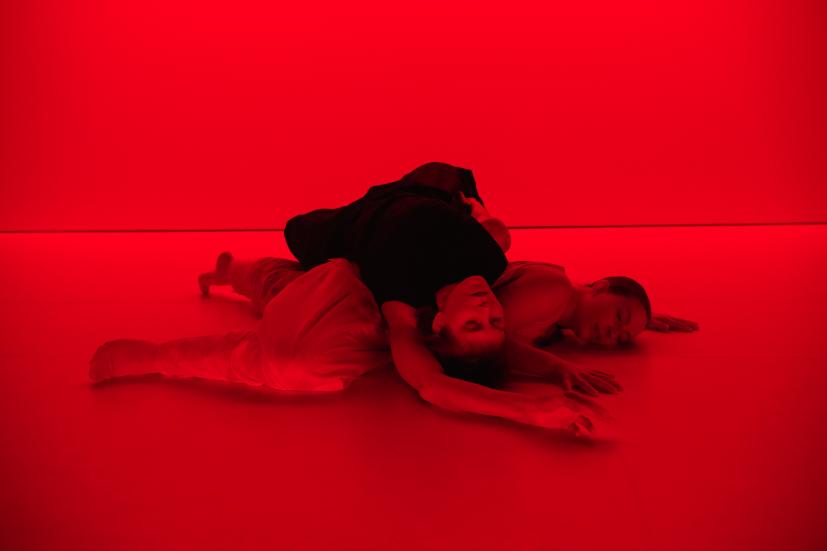 Kaksi tanssijaa makaa toistensa päällä lattialla. Kuva on hyvin punainen