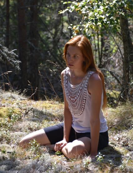 Matilda Aaltonen in a summery forest, sitting on the ground, she is looking down