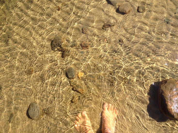 Picture of a clear beach water, the picture is taken above the water and there is two feet in the water