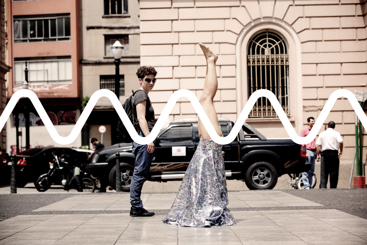 Photo from a street. One person is standing on their head. Their head is covere my a skirt. Another person is looking at them.