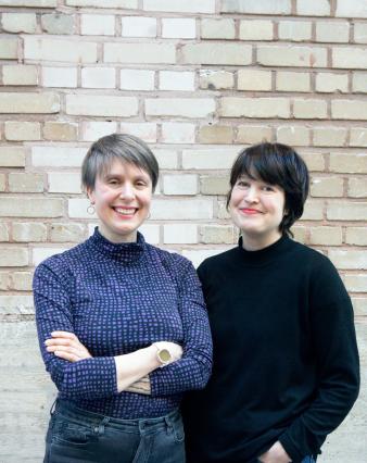 Milla Martikainen & Kati Raatikainen stading in front of a brick wall, smiling.