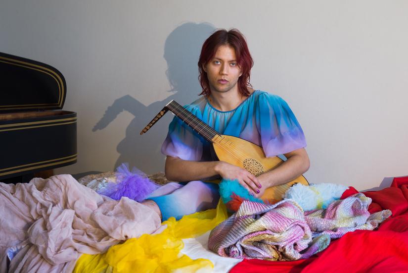 A person is sitting in a pile of colourful clothes. They are holding a lute.