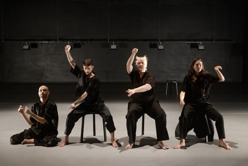 Neljä mustiin pukeutunutta esiintyjää. Kolme istuu tuolilla, yksi lattialla.