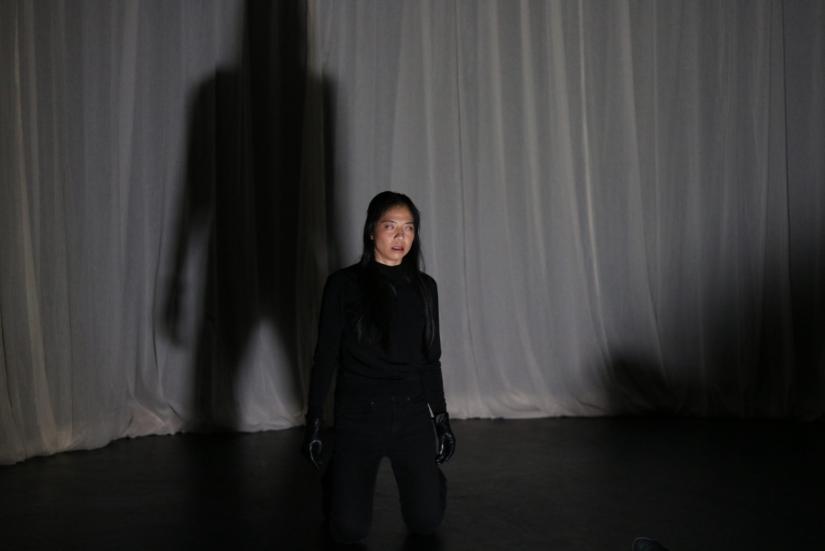 Dancer standing in a black overall suit in the centre in front of a white curtain. Dancer has her Eyes semi-closed