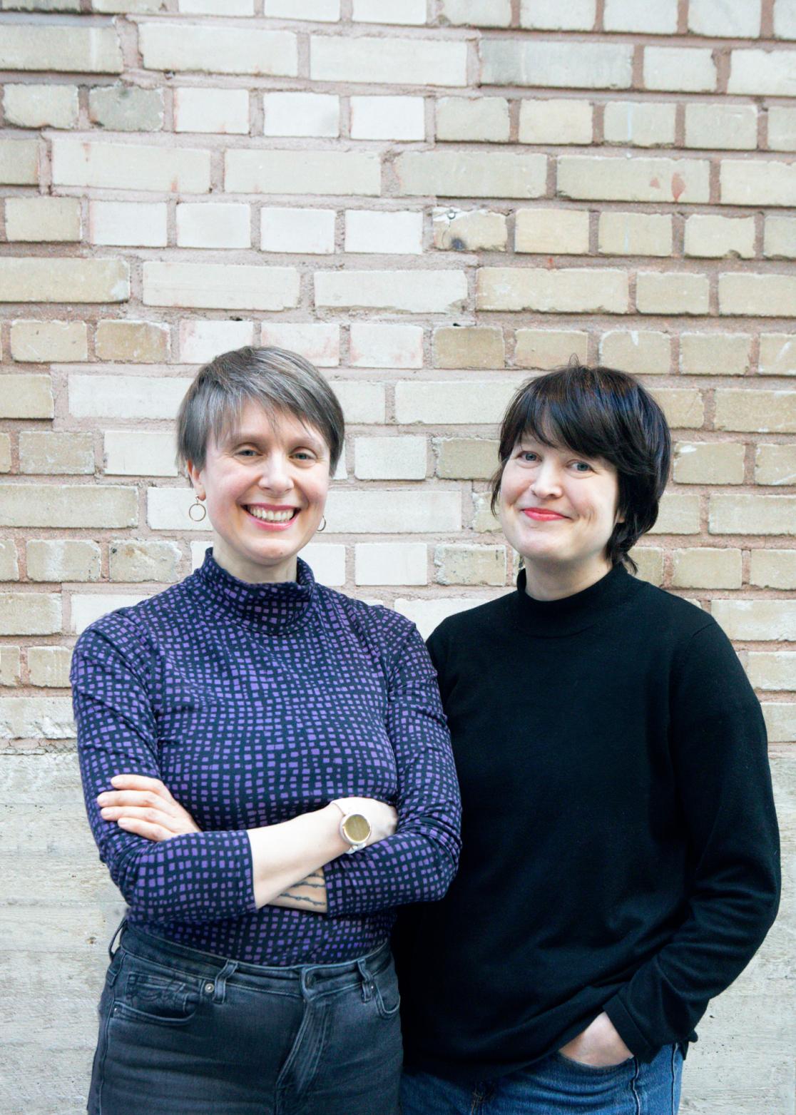 Milla Martikainen & Kati Raatikainen stading in front of a brick wall, smiling.