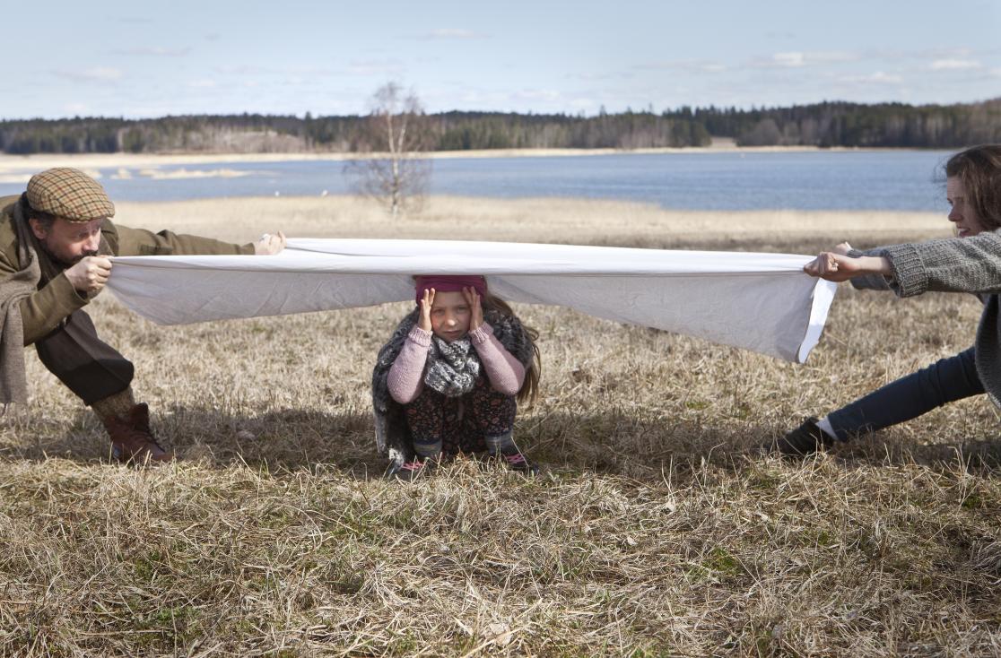 Kaksi aikuista ja yksi lapi ulkona. Aikuiset vetävät lakanaa, jonka alla lapsi kyykyssä.