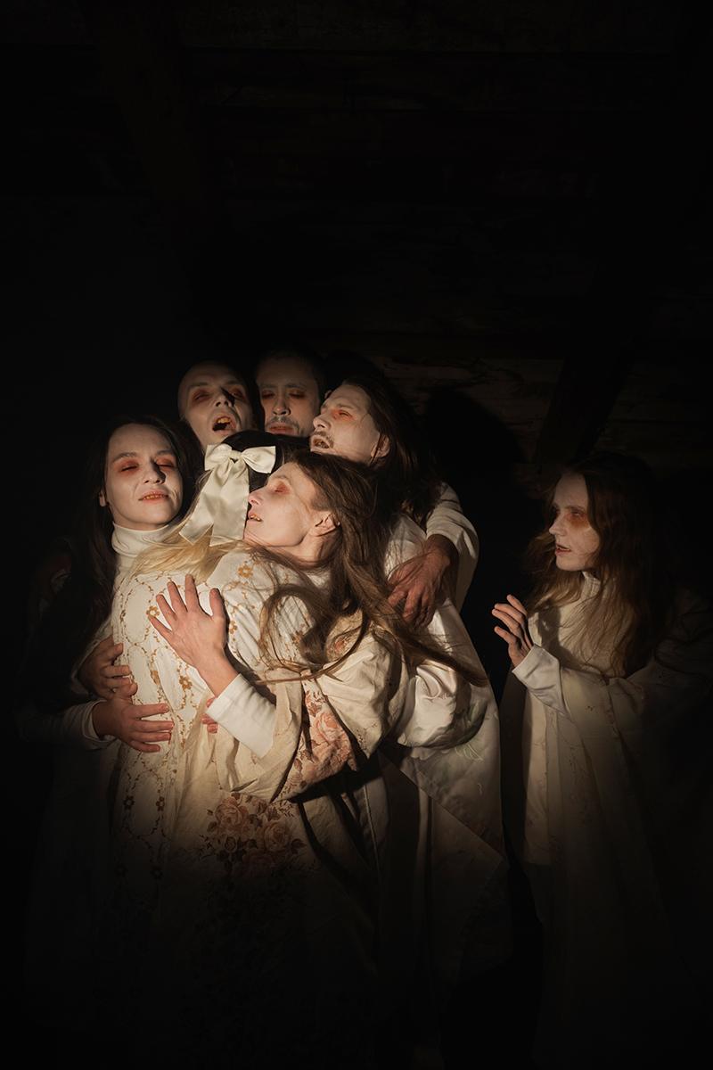 Six dancers in white gowns in a soft spotlight hugging one dancer who has their back to the camera