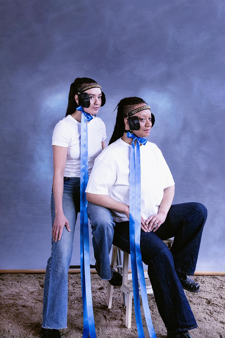 Artists Marika Peura & Caroline Suinner pose in front of blue background in white shirts and jeans, wearing horse brindles with blinkers and blue bows.