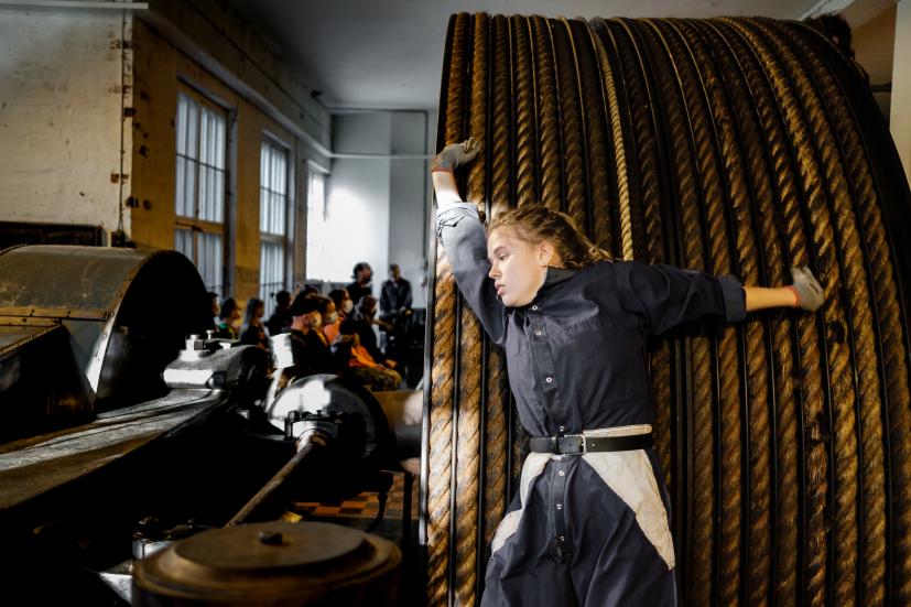 Performer is leaning against a big reel of rope. Audience at the background.