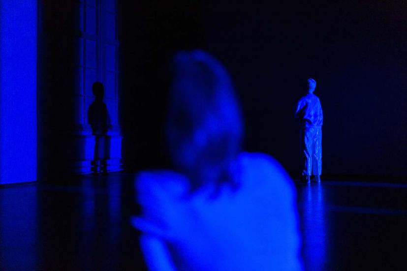 A space with deep blue light. One dancer is close to the camera, with the back turned. The other dancer can be seen in the background.