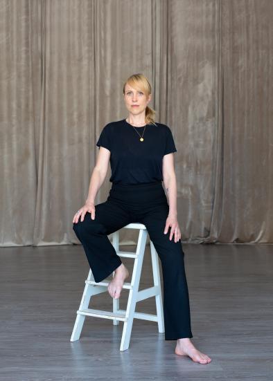 Anni Rissanen mustissa vaatteissa puoli-istuvassa asennossa valkoisen tuolin päällä.