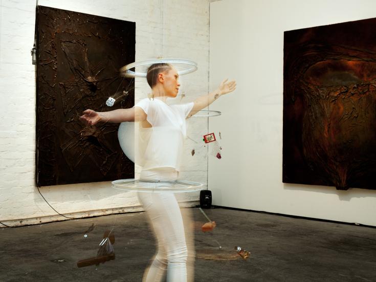 Anni Rissanen valkoisissa vaatteissa, kädet ojennettuina sivuilla, tekemässä pyörivää liikettä, galleriatilassa.
