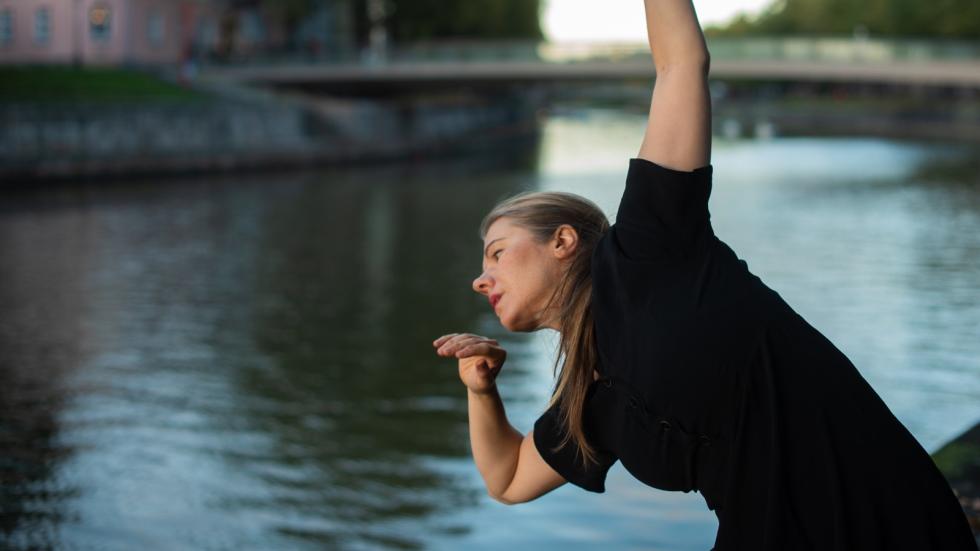 Maria Nurmela sivulle taipuneessa asennossa kädet ylhäälle osoittaen joen rannassa tanssimassa.