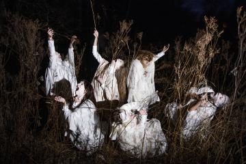 Six people in white ghost-like outfits standing in a dried up bush in the dar, with a spotlight on them