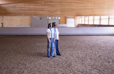 Artists Marika Peura & Caroline Suinner stand in the middle of a riding hall in white shirts and jeans, wearing horse brindles with blinkers and blue bows.