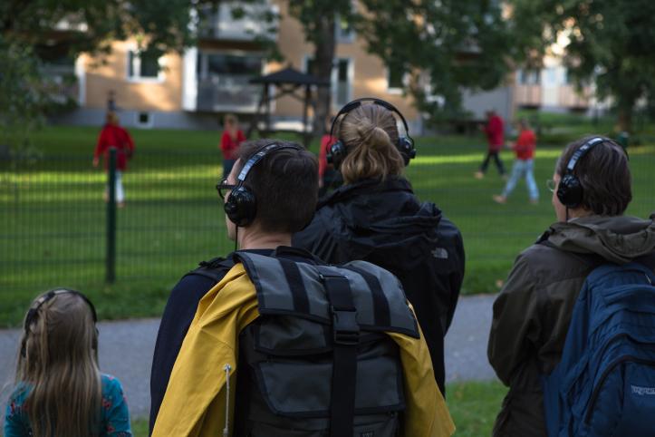 Kuulokkeet päässä seisovat ihmiset katsovat aidan toisella puolella tanssivia henkilöitä.