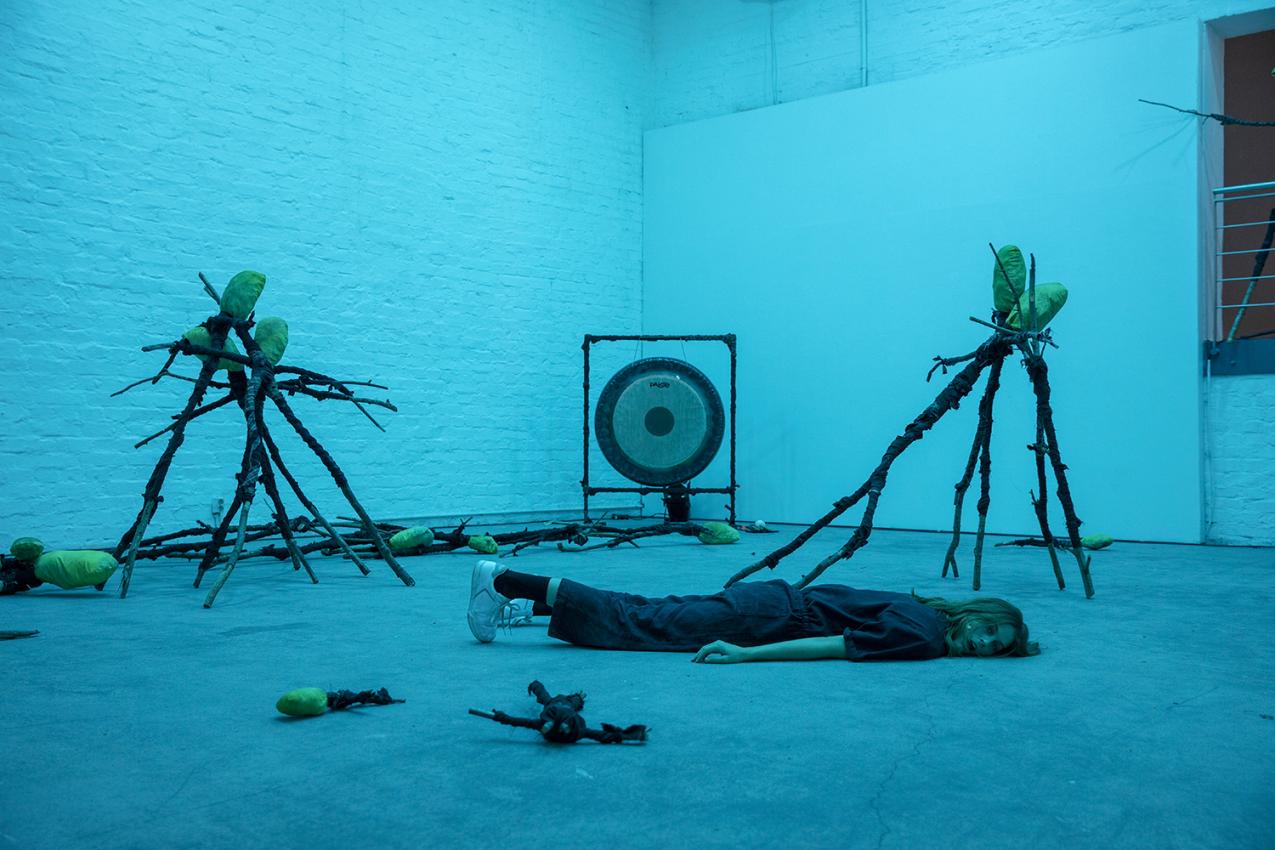A blue-tinted room. One performer is lying on the floor on their stomach. There are some twigs and a cong in the space.