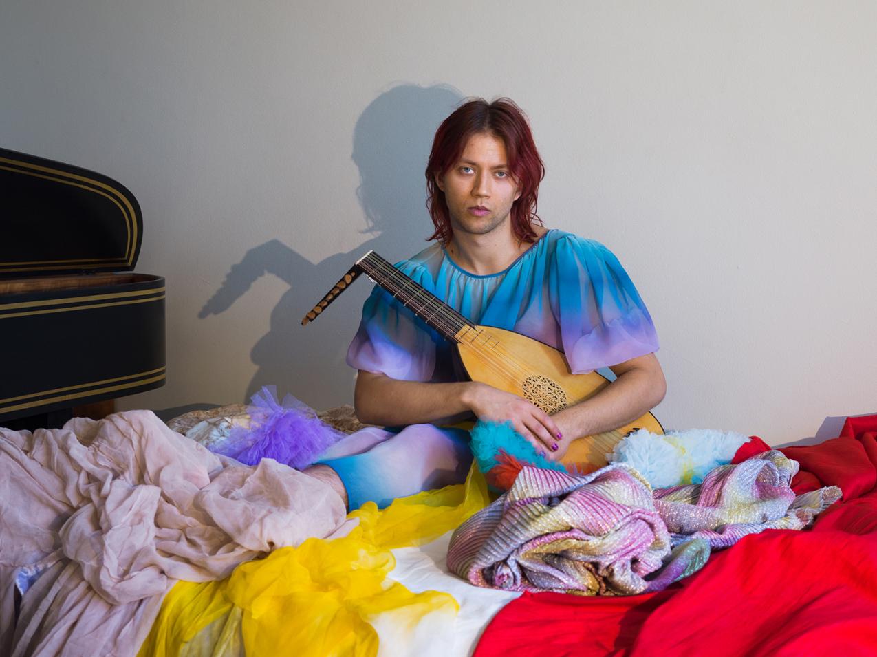 A person is sitting in a pile of colourful clothes. They are holding a lute.