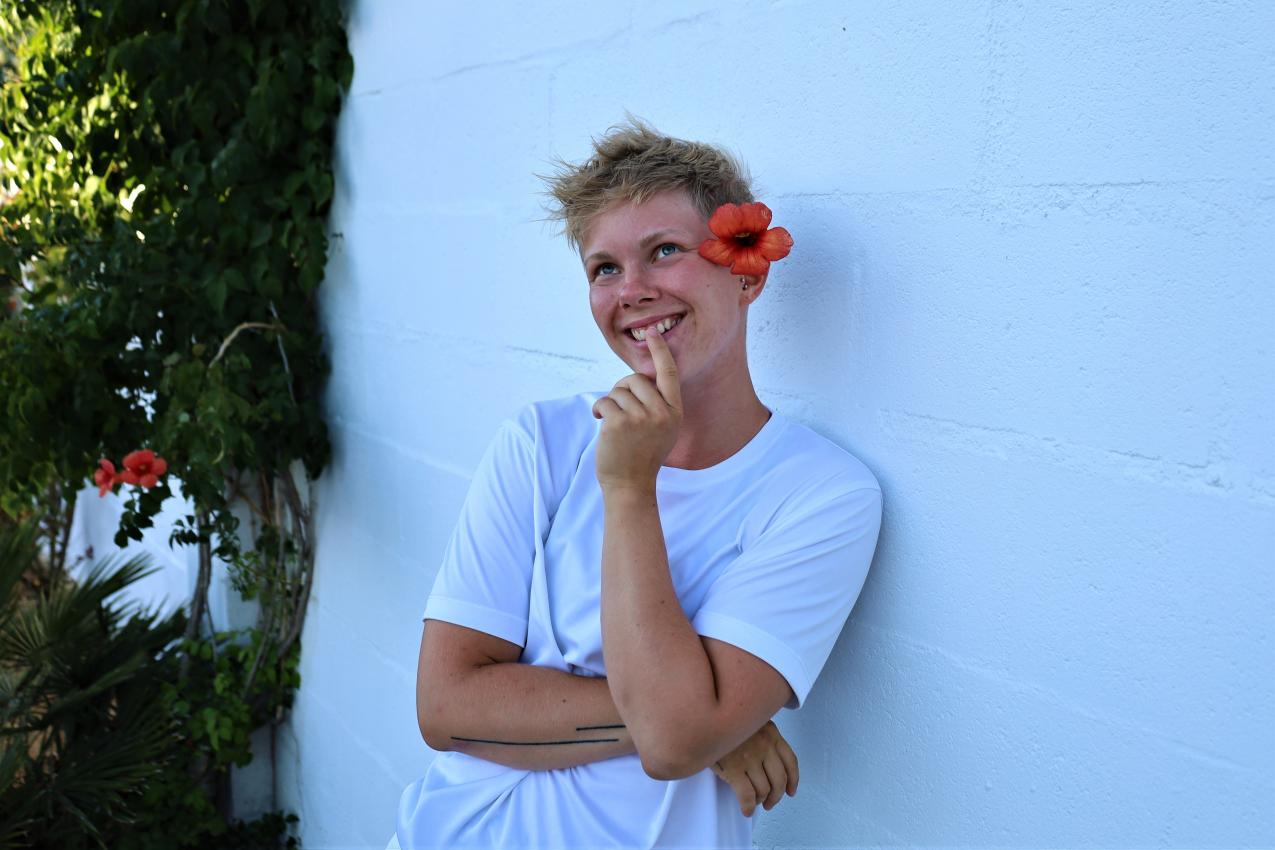 A person in a white t-shirt is leaning against a wall, smiling. They have a flower behind their ear.