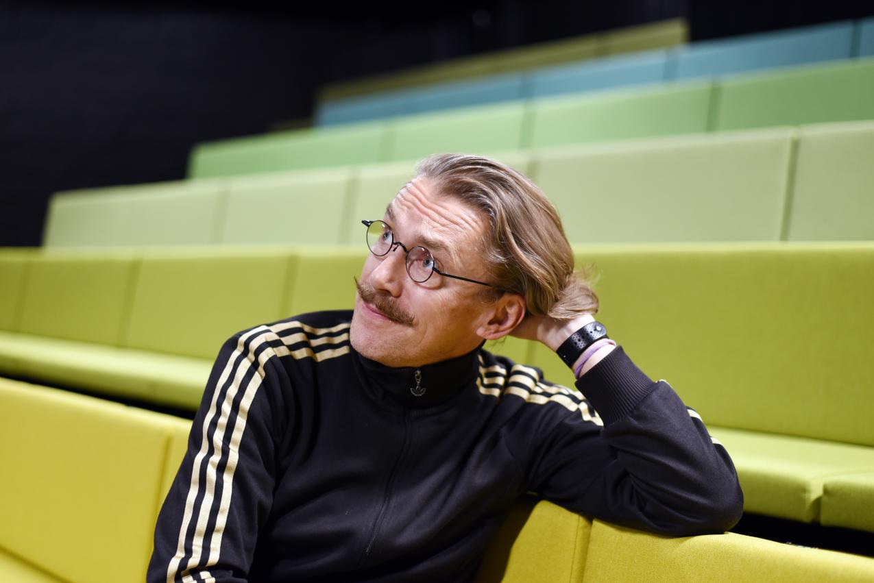 Jyrki Karttunen istumassa teatterin katsomossa, jossa on keltavihreät penkit. Hän nojaa kyynärpäällään selkänojaan ja katsoo oikealle yläviistoon.