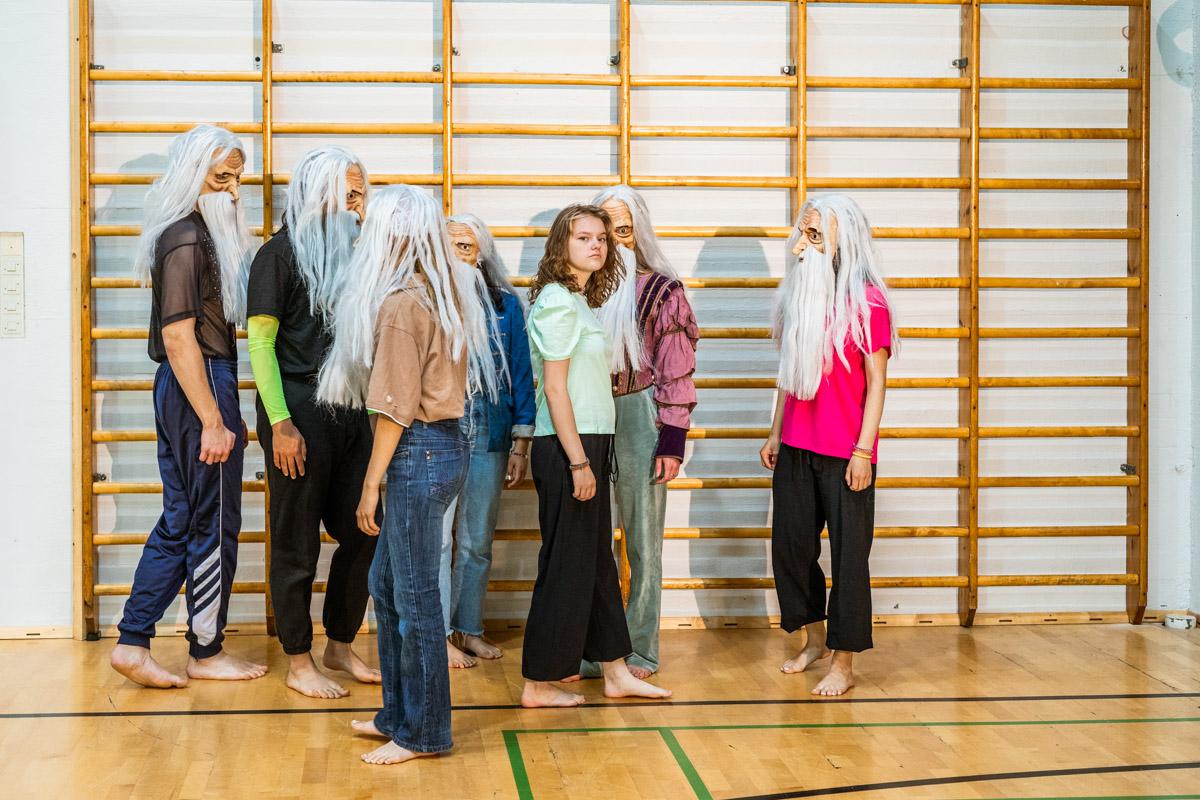 Ryhmä nuoria seisoo koulun liikuntasalissa. Yhtä lukuunottamatta heillä on päässään naamiot. Naamiot esittävät vanhaa miestä, jolla on pitkä valkoinen parta ja tukka.
