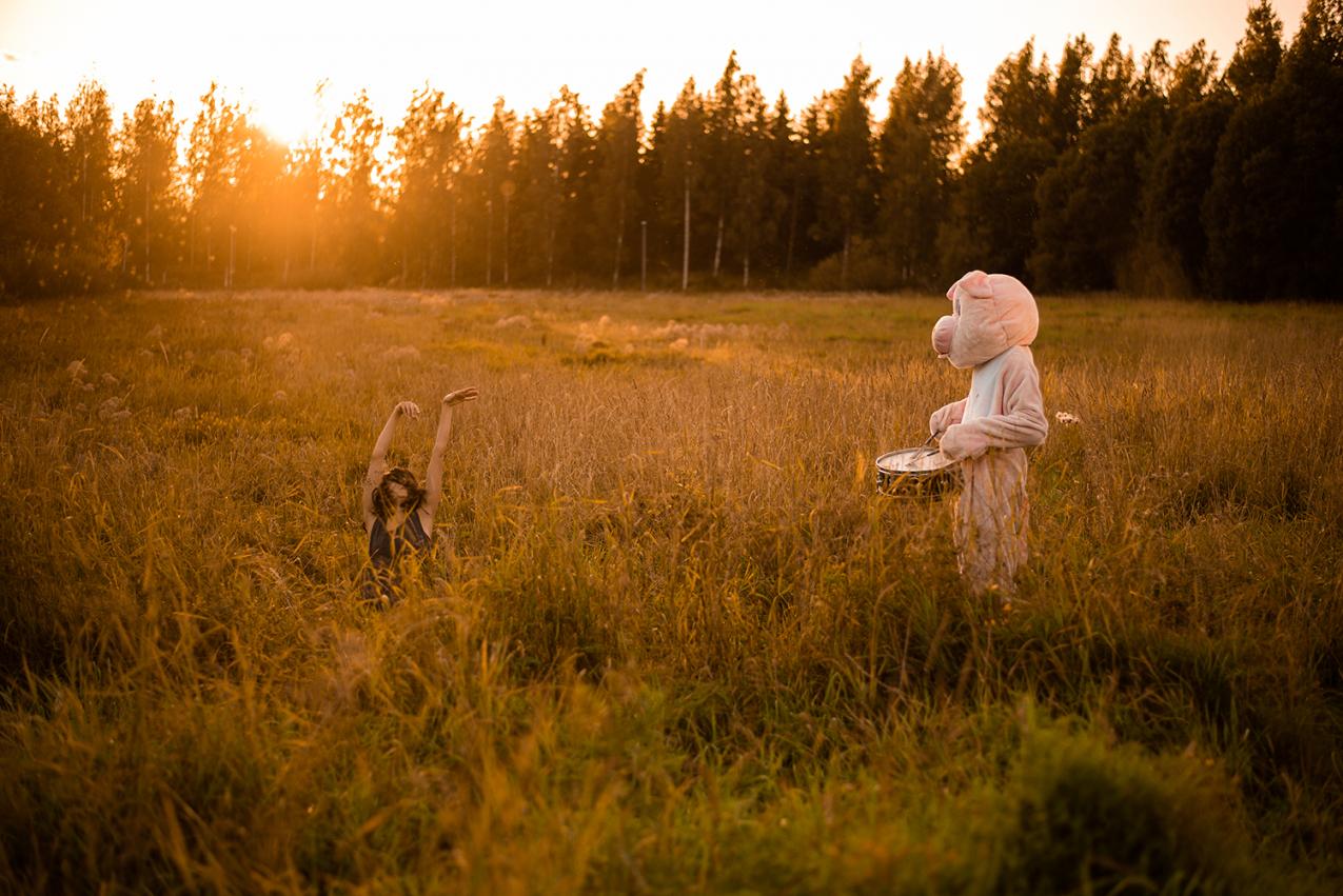 Tanssija ja possupukuun pukeutunut esiintyjä niityllä ilta-auringossa