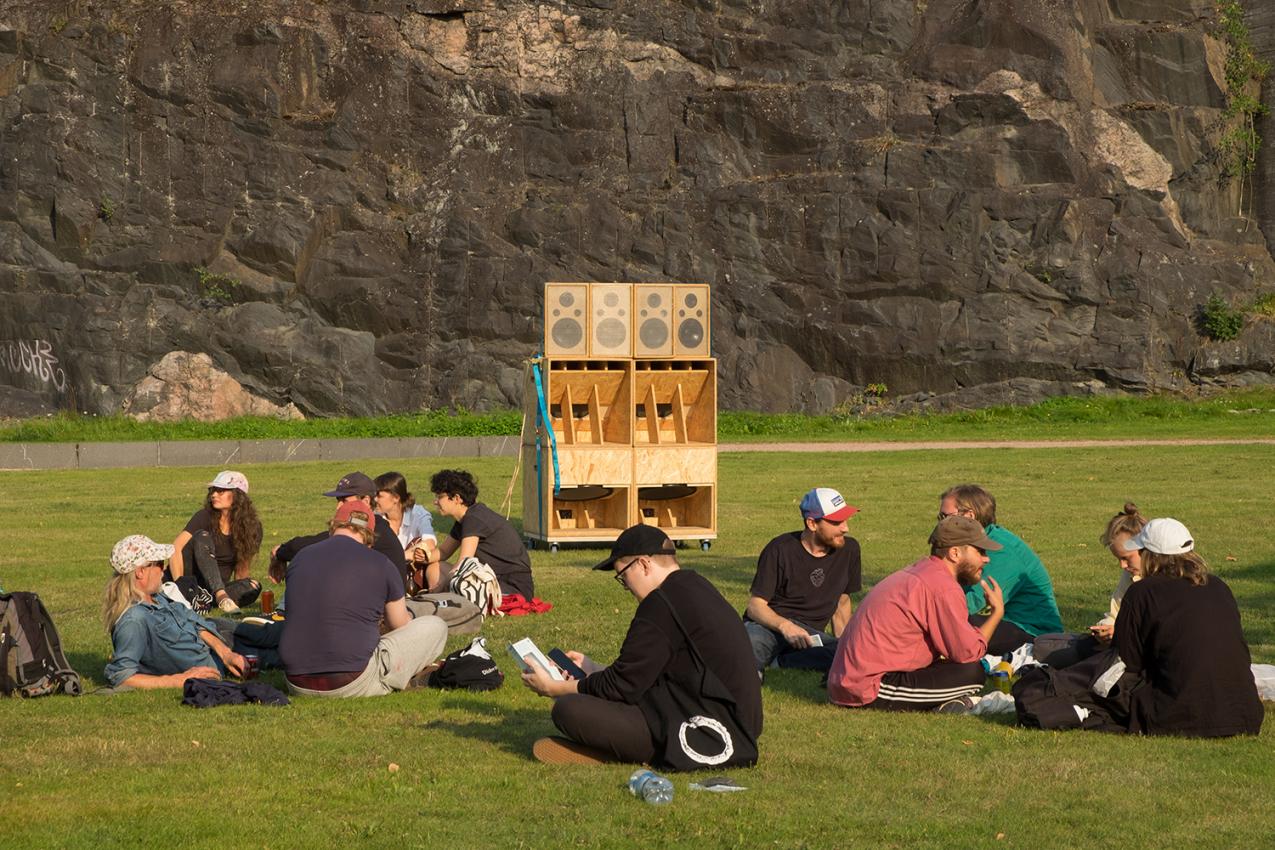 Several people sitting on grass, as if on picnic. There is a wooden sculpture in the middle of the photo