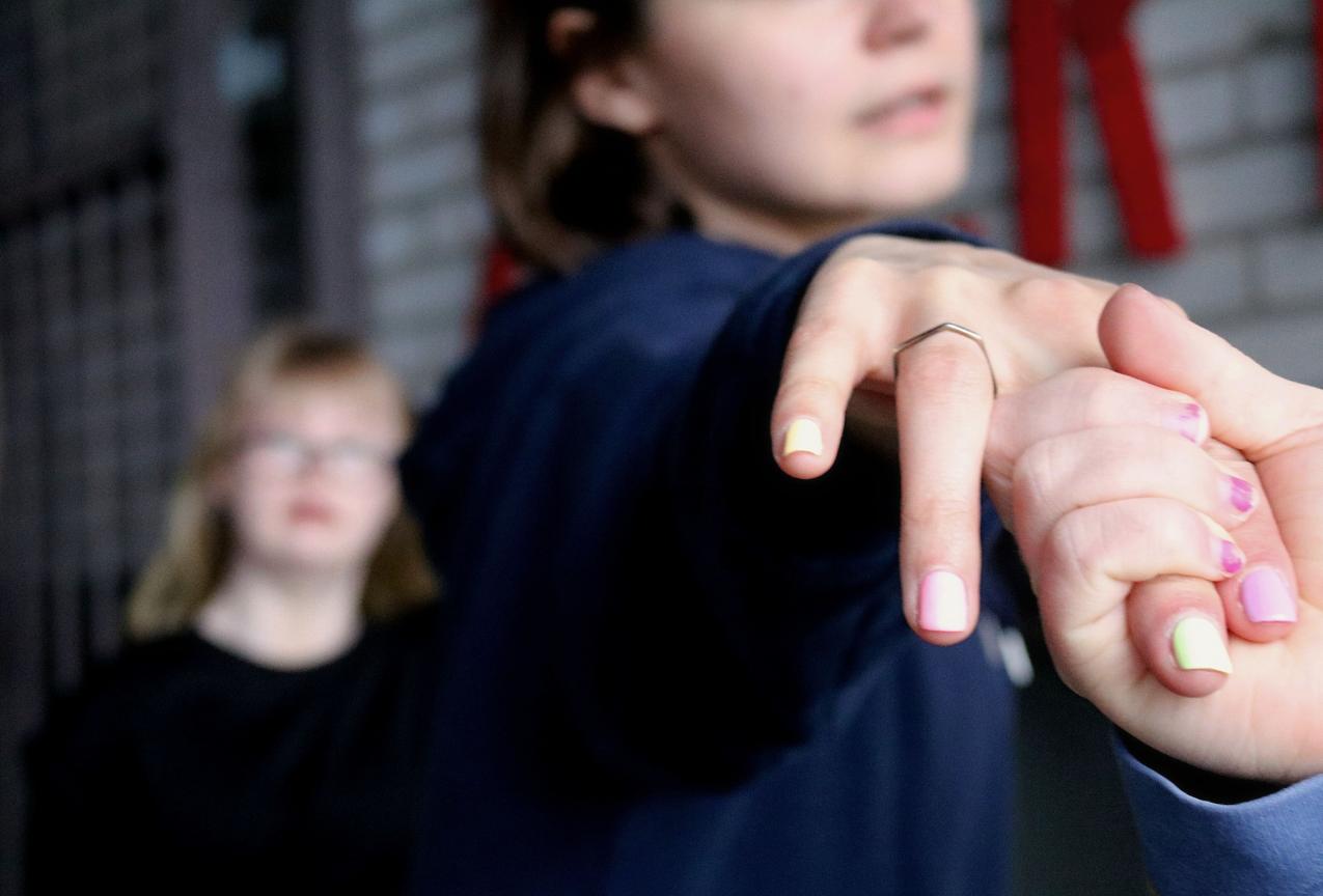 Hand in hand. Both hands have colorful nail polish on and on the other hand there is a ring. In the background there are two blurry human figures. The background is dark.