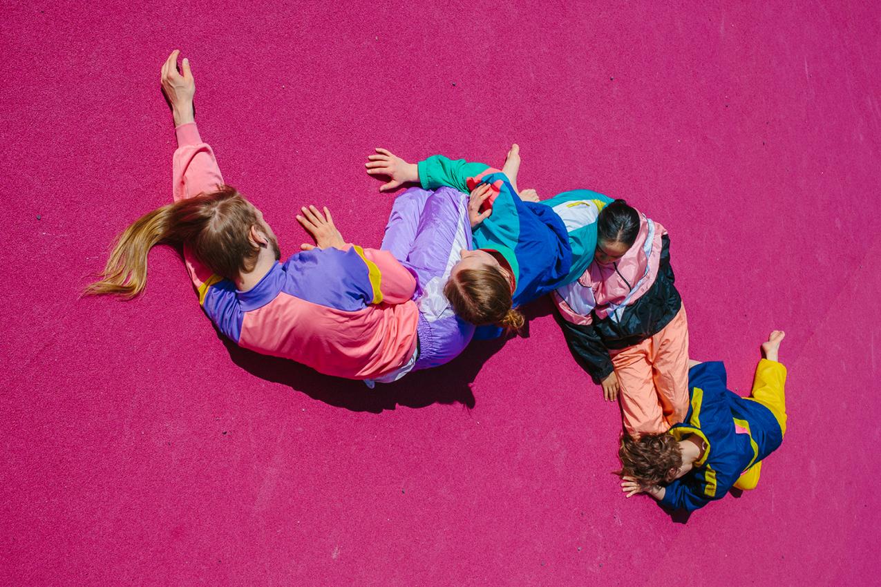 People in colourful clothes are lying on the gorund. They form a serpent like form on the bright pink ground.