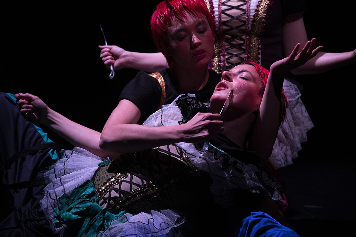 Three performers in a dimly lit space. They are lying on top of each other
