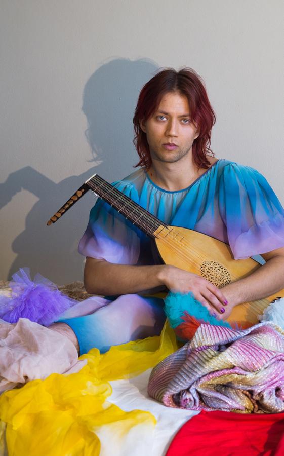 A person is sitting in a pile of colourful clothes. They are holding a lute.