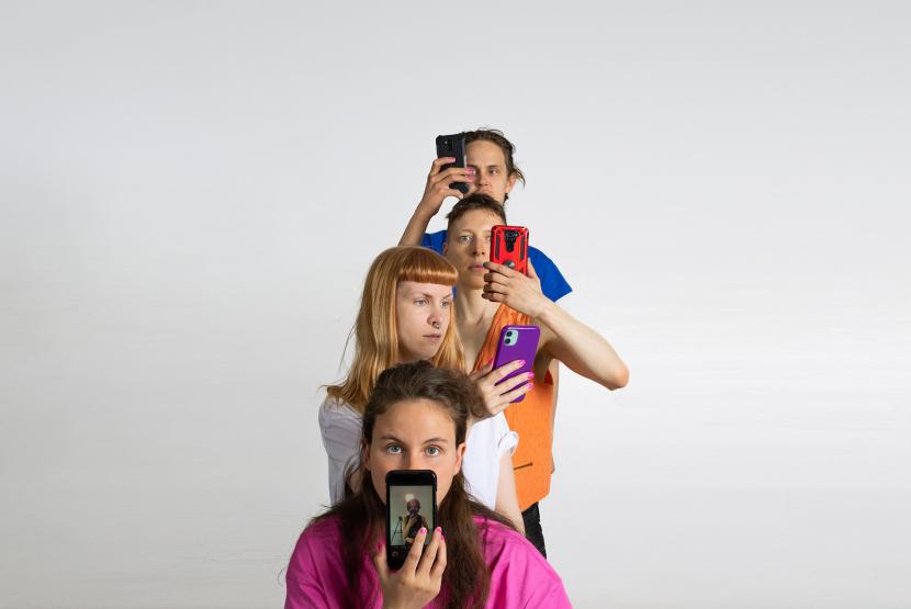 Four people are standin in a row. They are facing the camera. Each is holding a phone in their hand.