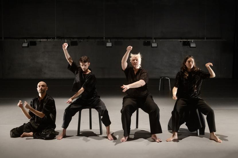 Neljä mustiin pukeutunutta esiintyjää. Kolme istuu tuolilla, yksi lattialla.