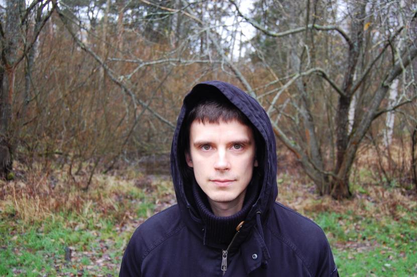 Mikko Niemistö, pukeutuneena mustaan huppariin, katsoo kameraan taustalla metsä