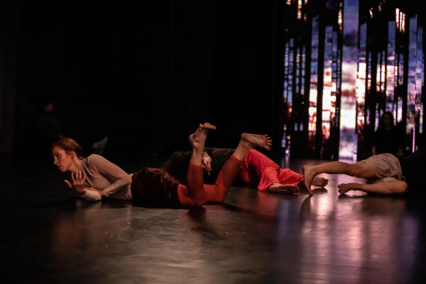 Perfomance picture of a darkish space, where three people are lying on the floor in spiraly positions, Matilda Aaltonen is in the front.