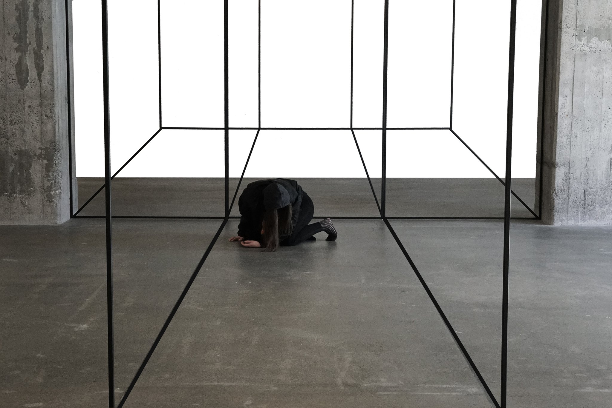 A figure crouch on the floor in a gallery like space, which also has metallic frames in it.