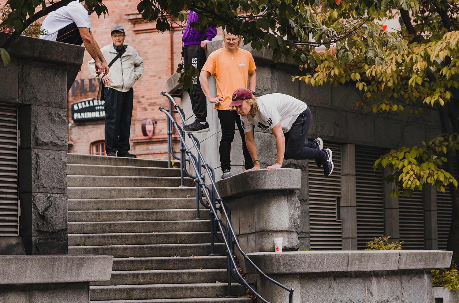 A staircase in a publci space. One person is jumping over the stony handrail, three other people are watching them.