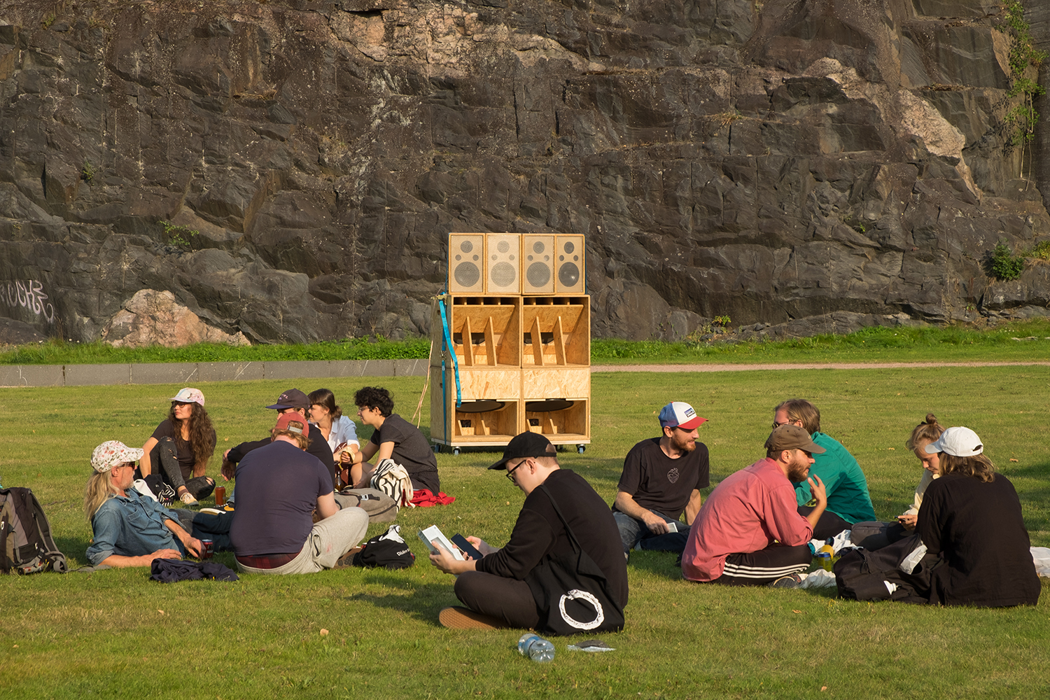 Several people sitting on grass, as if on picnic. There is a wooden sculpture in the middle of the photo